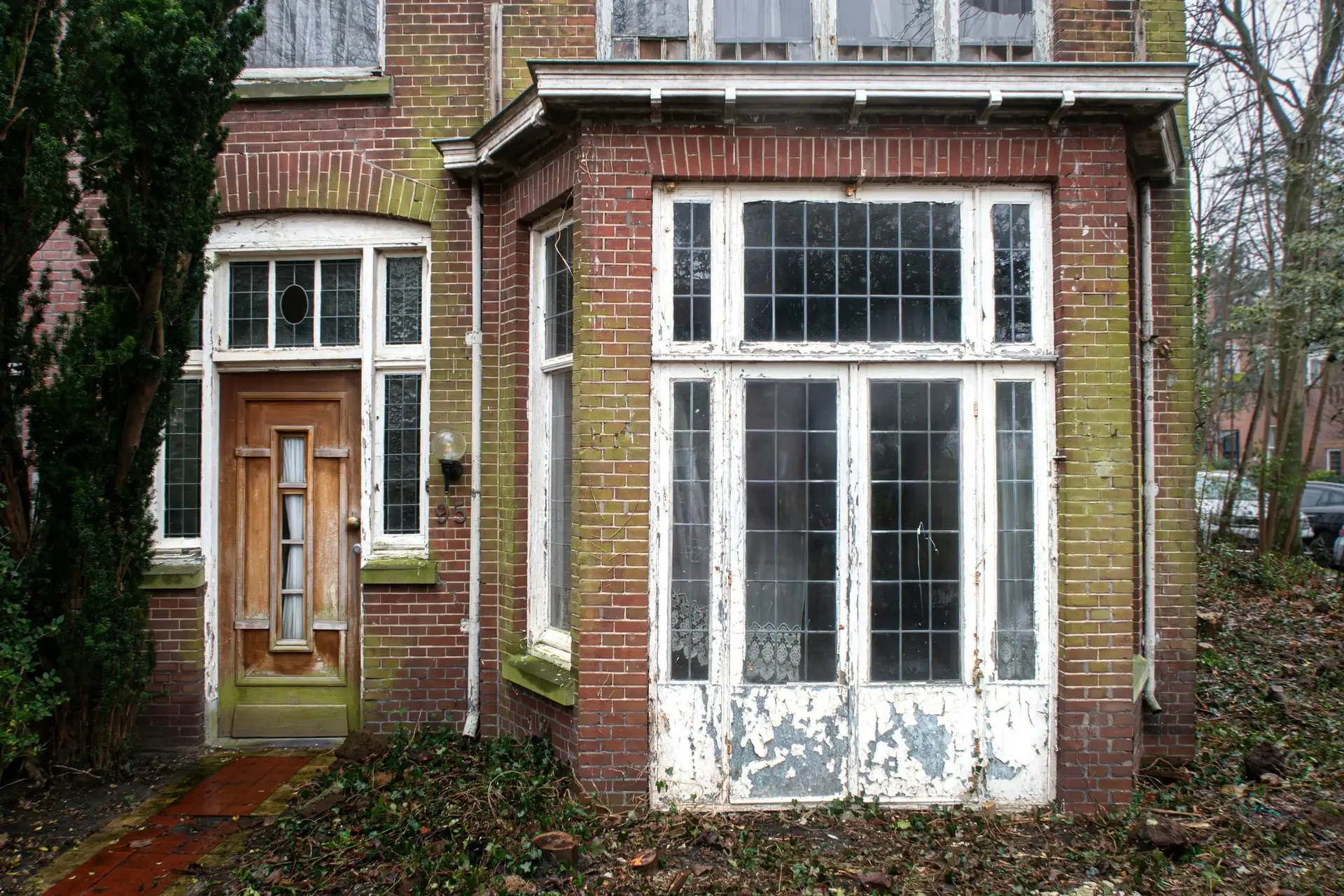 Oudere bakstenen woning met verweerde witte ramen en een verwaarloosde voortuin